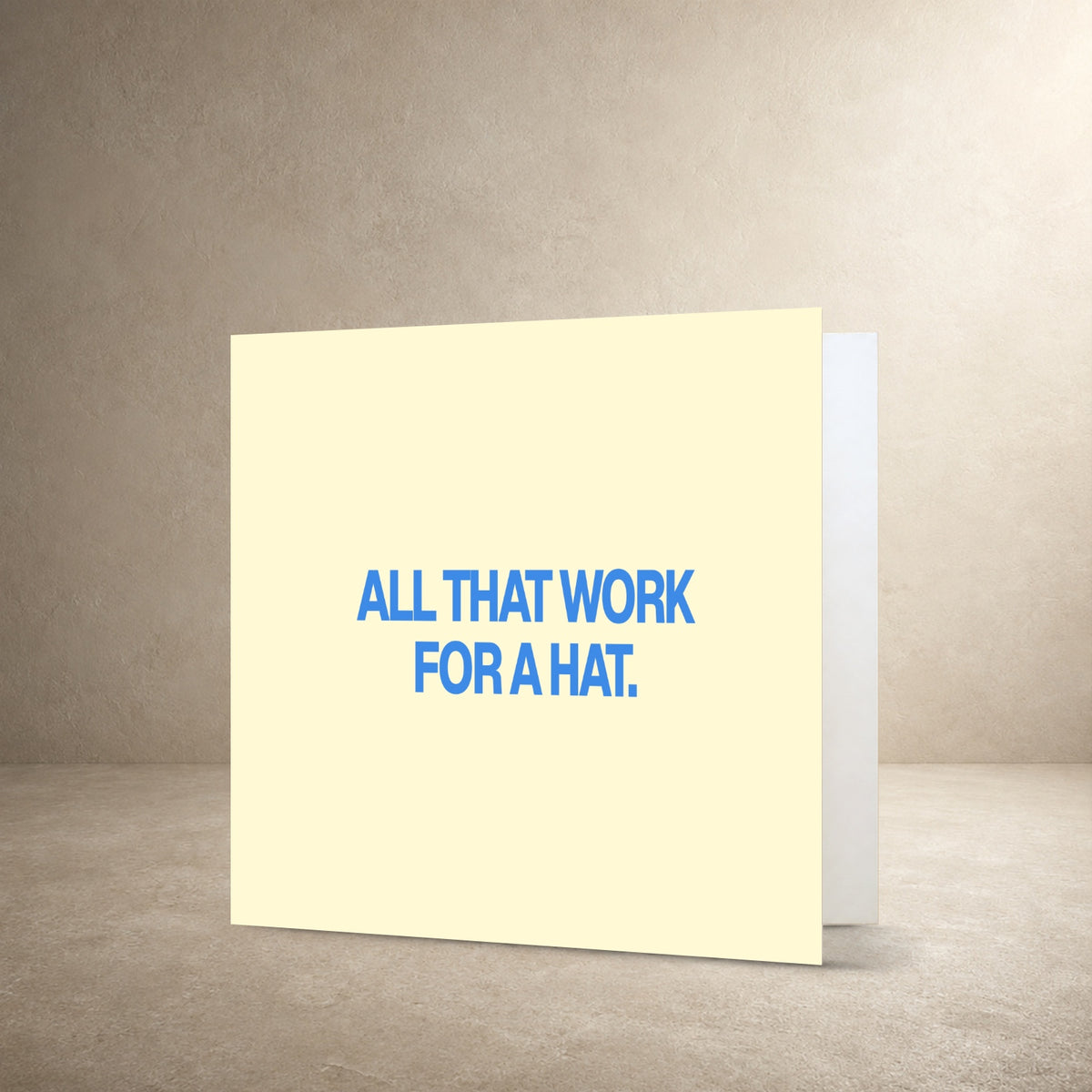 Pale cream card front with bold blue centred typography, with the message: “All that work for a hat.”.