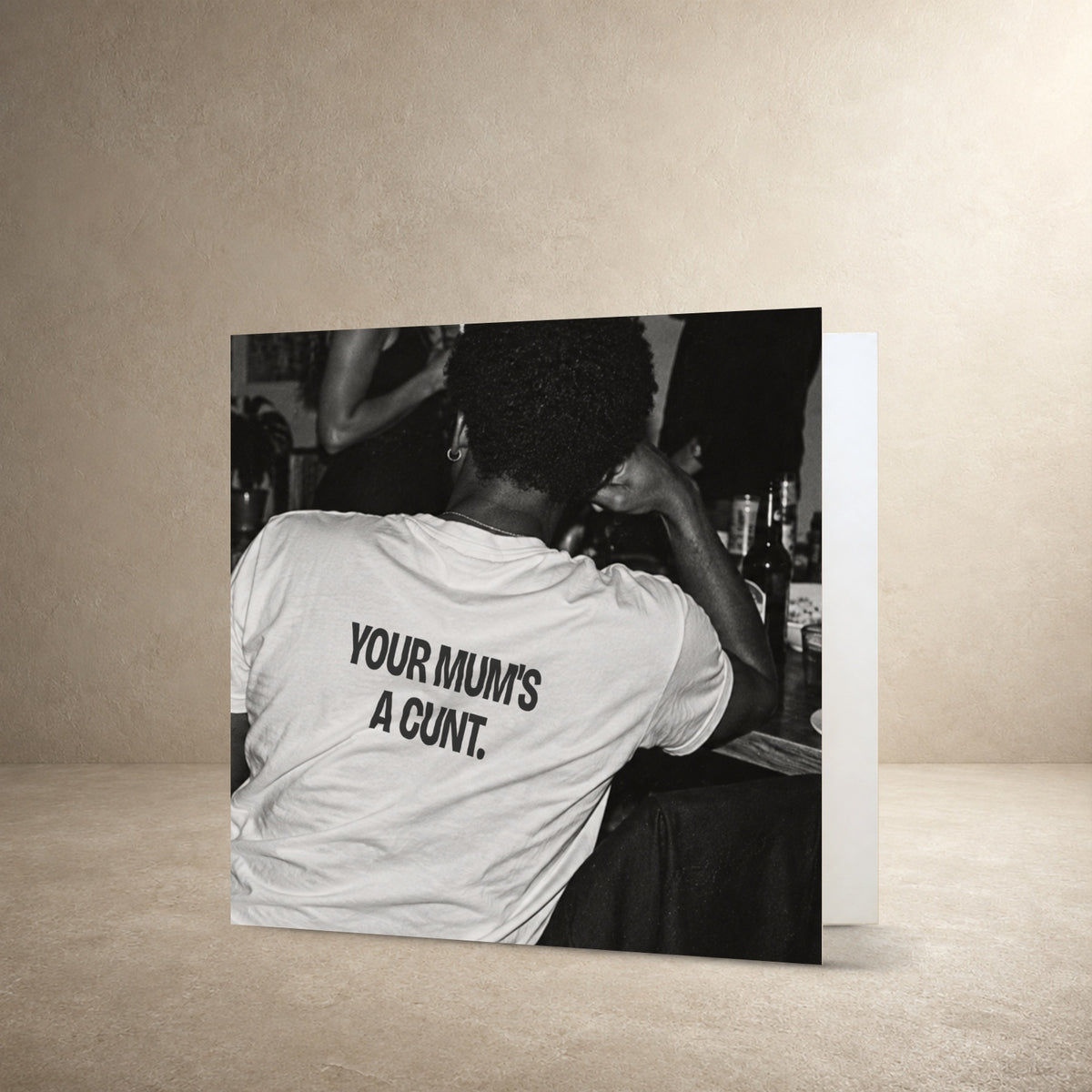 Black-and-white rear-view photograph of a person wearing a white T-shirt, with the message: “Your mum's a cunt.”.