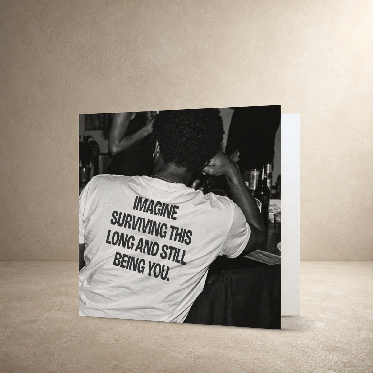 Black-and-white rear-view photograph of a person wearing a white T-shirt, with the message: “Imagine surviving this long and still being you.”.